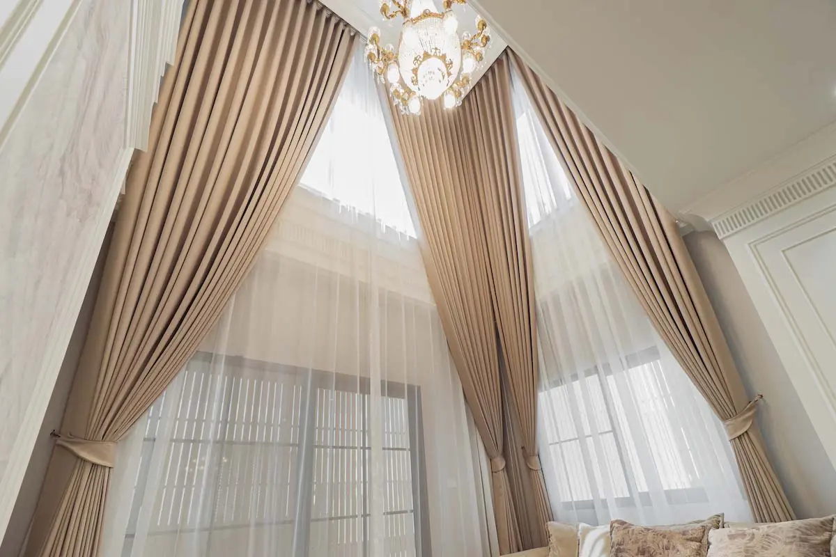 Tall beige curtains with sheer white layers, elegantly draped in a high-ceiling room and a classic chandelier.