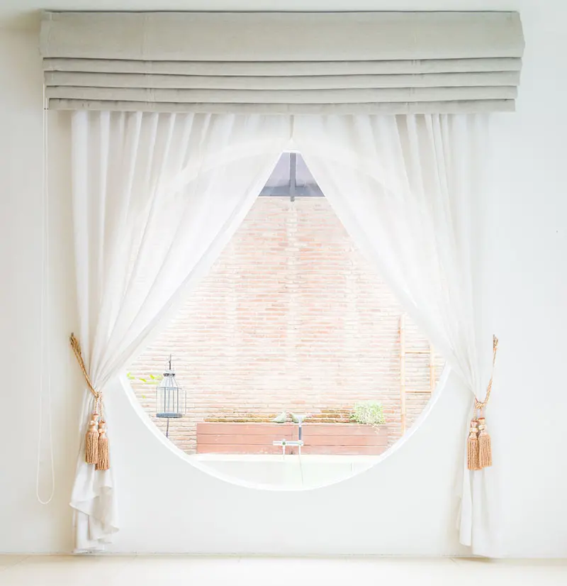 Circular window adorned with side-tied white sheer curtains and a raised Roman shade as the secondary layer.