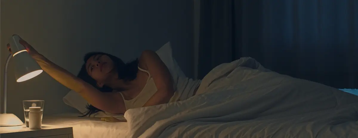 A girl lying in bed reaches out to turn off the night lamp situated next to her bed.