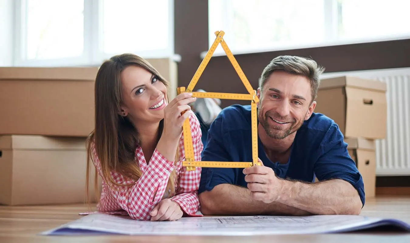 A couple holding a wooden house frame smiles while lying on blueprints.