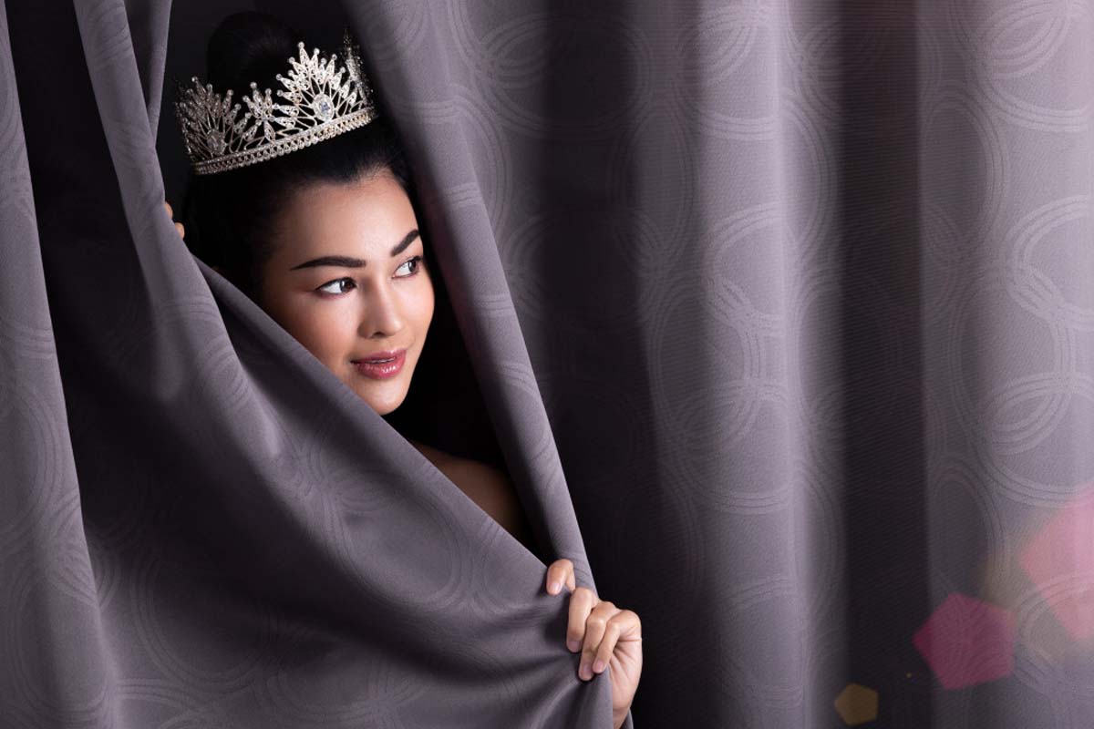 Woman with a tiara peeking through elegant grey night curtains.