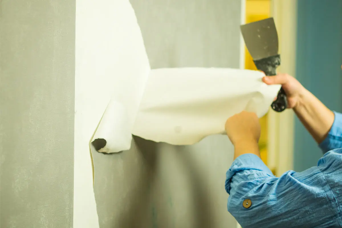 Person in a blue shirt removing old wallpaper with a scraper, revealing a bare wall underneath.