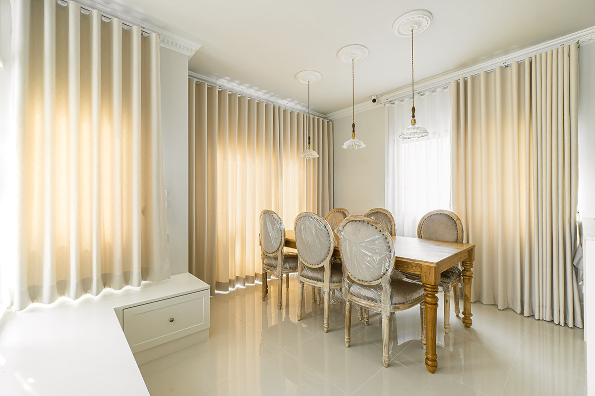 Dining area with cream dimout curtains, a wooden table, and upholstered chairs.