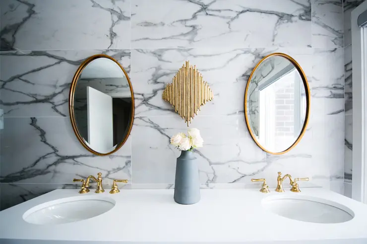 A bathroom with dual sinks and mirrors, earth-toned tiles, and gold-colored taps.