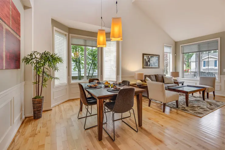 A living room in earthy hue with a dining area, seating section, and three windows adorned with blinds.