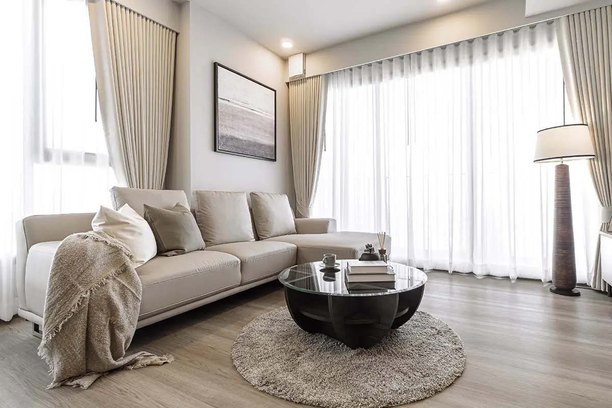 Modern living room with light cream curtains, a cozy beige sofa, and a round glass coffee table.